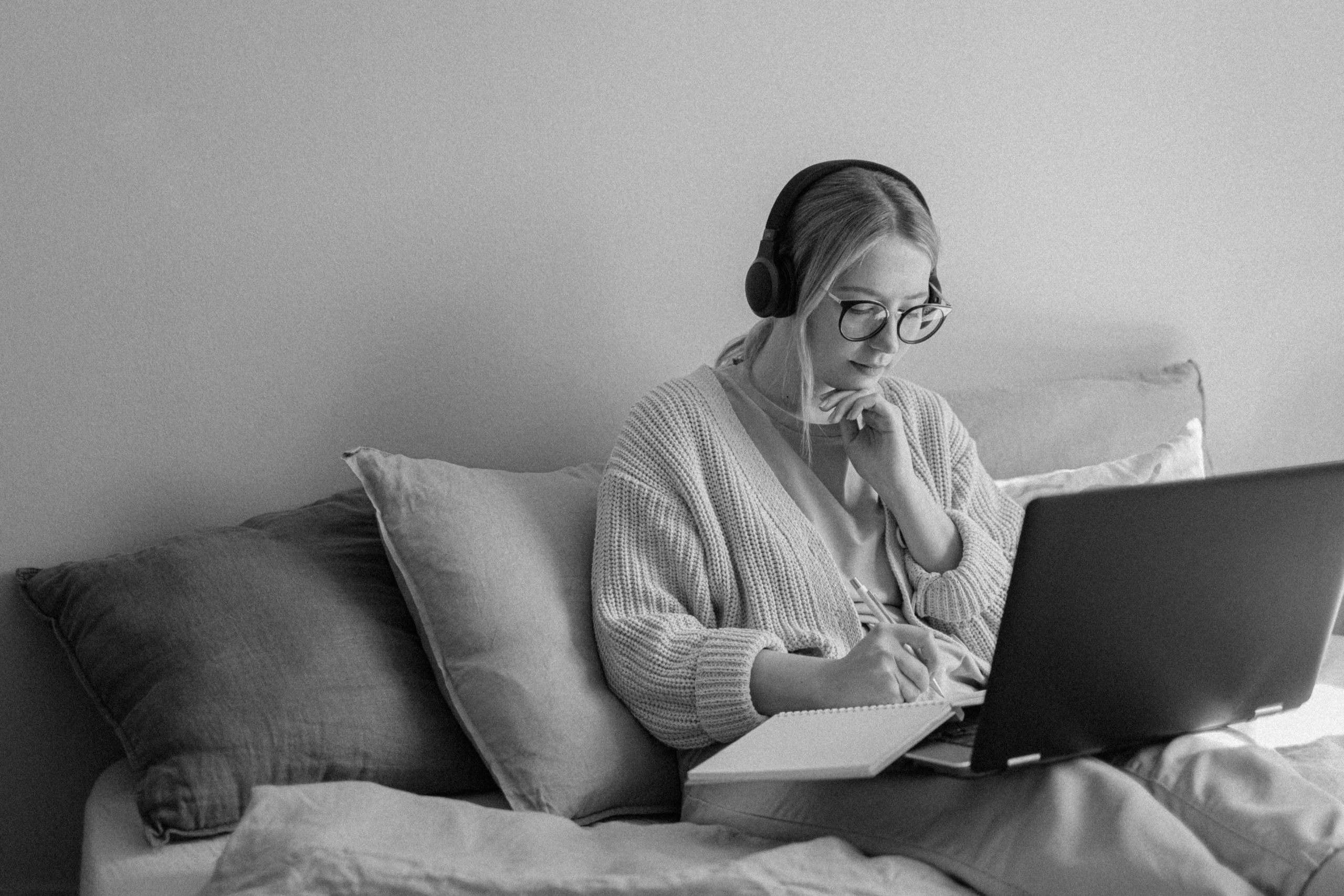Young woman sitting on her couch, wearing headphone and working on her laptop.